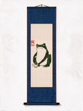 A vertical Japanese-style hanging scroll artwork on a white background. The scroll has a dark blue border and a central beige panel featuring a green frog illustration with a small red seal.