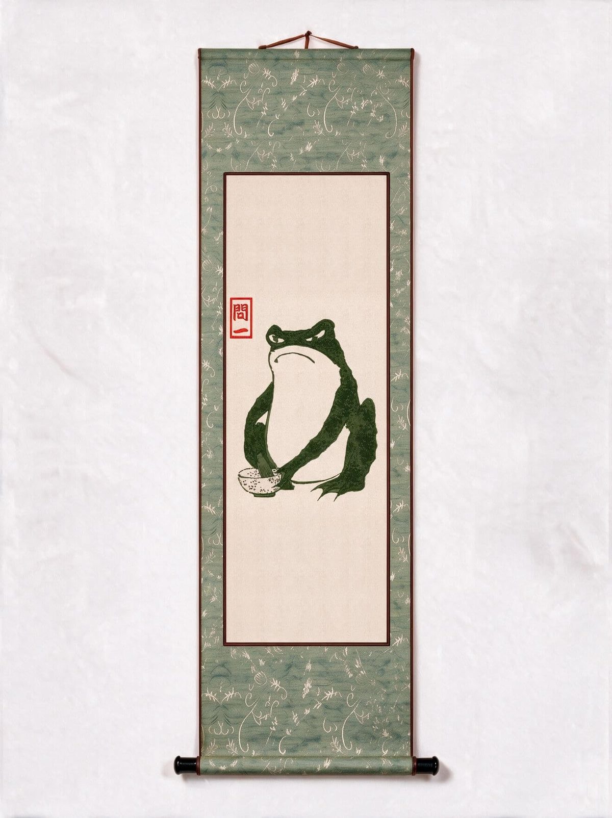 A traditional Japanese hanging scroll wall art featuring a green frog illustration on a beige center panel, bordered by a textured sage green patterned fabric with a red seal.