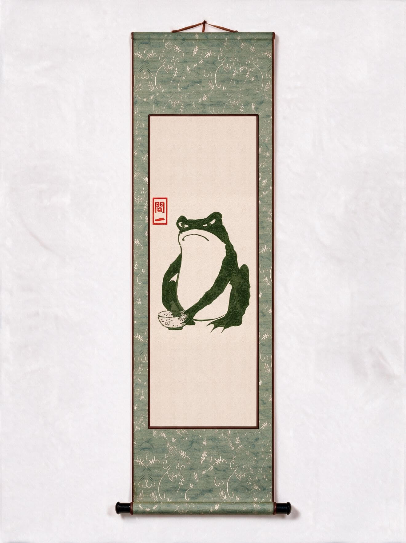 A traditional Japanese hanging scroll wall art featuring a green frog illustration on a beige center panel, bordered by a textured sage green patterned fabric with a red seal.