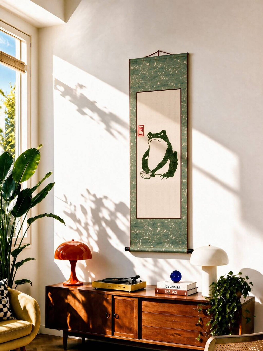 A vertical Japanese-style hanging scroll artwork featuring a green frog illustration on a cream center panel, displayed on a white wall in a sunlit room above a wooden cabinet with plants and decor.